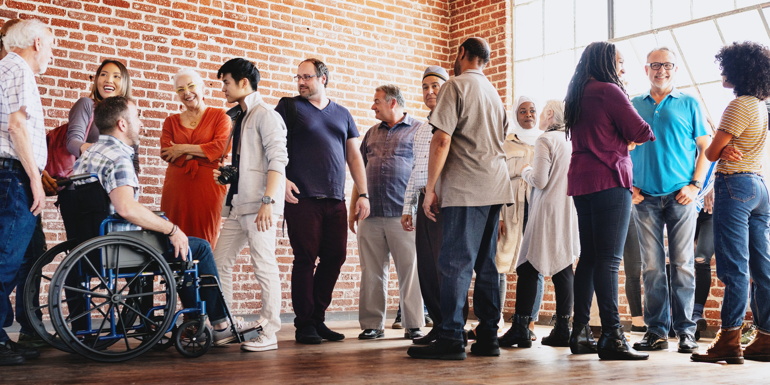 Diverse group of people socializing indoors. Men and women of various ethnicities and ages, including a person in a wheelchair, gathered in a brick-walled room.