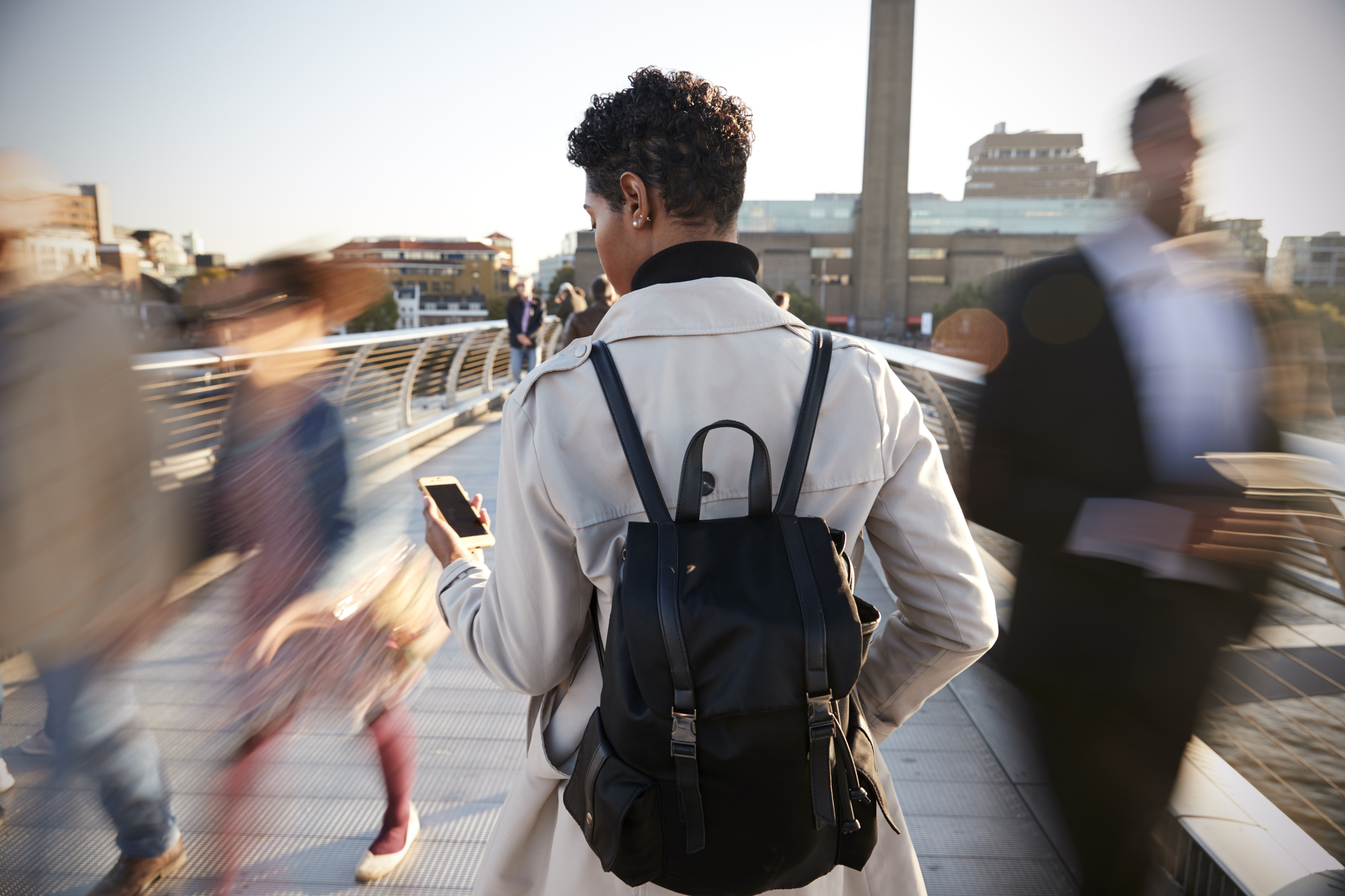 Back view of young adult woman standing on Millennium Bridge, London, using smartphone, motion blur